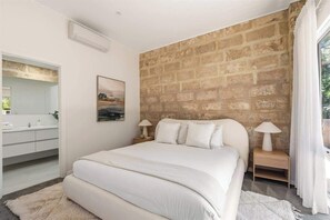 Ein Schlafzimmer mit Backsteinwand, einem Bett, Nachttischen inklusive Lampen, einem Gemälde und einem Badezimmer mit Spiegel und Waschbecken.