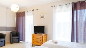 A hotel room with two beds, a TV on a wooden dresser, a chair, and a large window with curtains. - Cozy Studio with Private Terrace, Air Conditioning & Pool Table – Cazaubon (Cazaubon)
