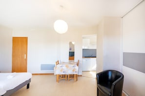 A room with a bed, a dining table with chairs, a black chair, and a kitchenette in the background. - Cosy Studio with Balcony, Air Conditioning & Pool Table – Cazaubon (Cazaubon)