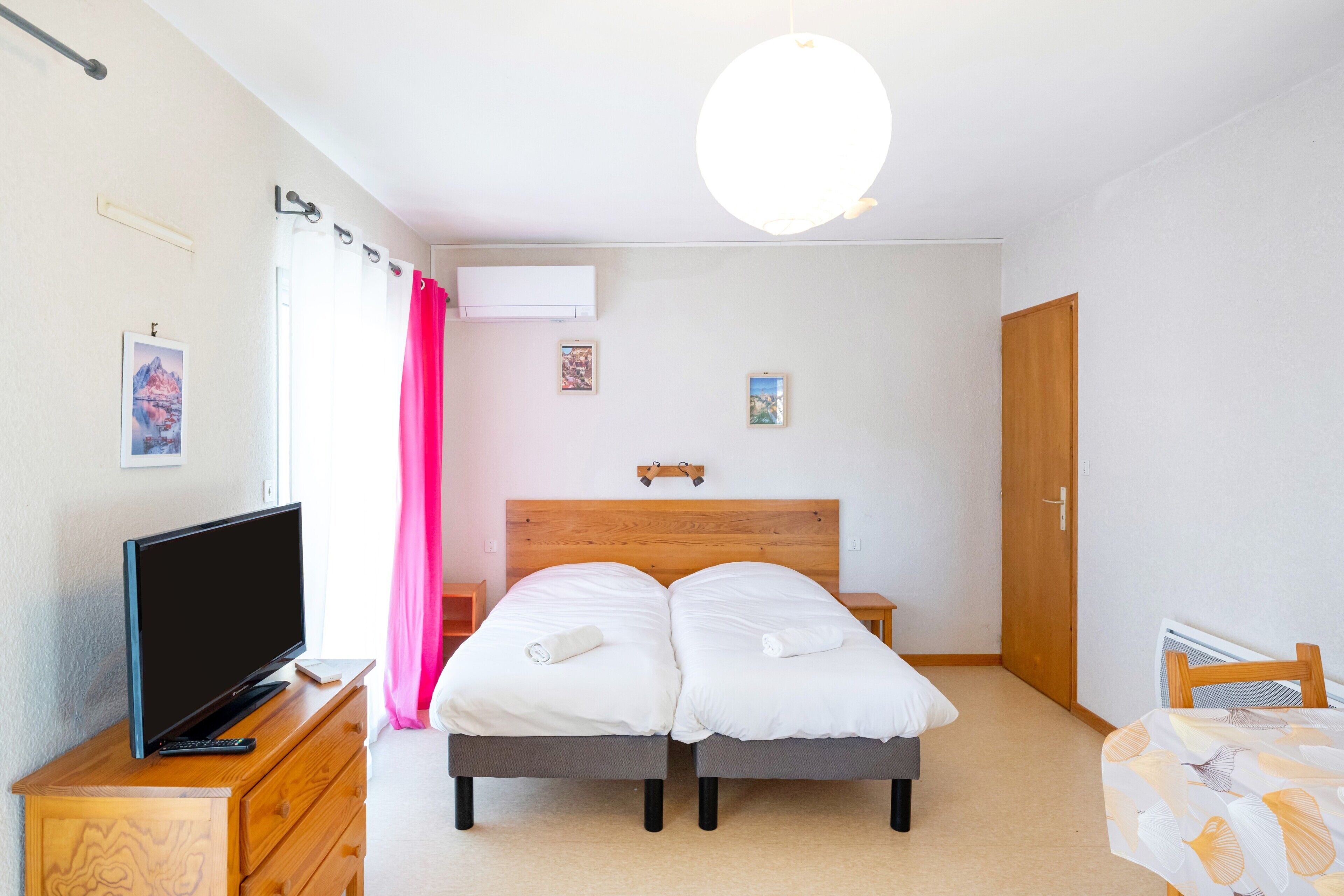 Ein Schlafzimmer mit einem Holzkopfteil, einem Fernseher auf einer Holzkommode, zwei Betten mit weißer Bettwäsche, einem rosa Vorhang und einer Deckenleuchte.