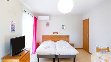 Ein Schlafzimmer mit einem Holzkopfteil, einem Fernseher auf einer Holzkommode, zwei Betten mit weißer Bettwäsche, einem rosa Vorhang und einer Deckenleuchte.