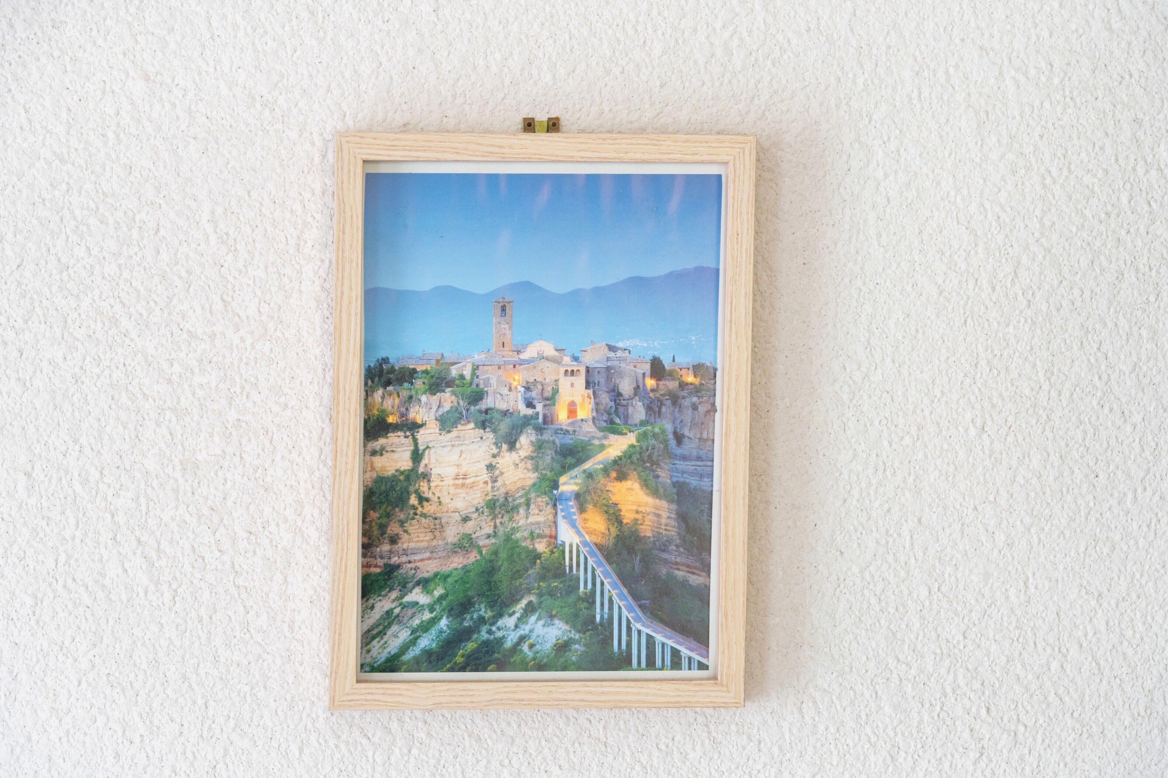 A framed painting of a mountainous landscape with a bridge and a tower.