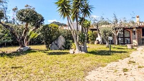 Un chemin de pierres mène à un bâtiment rustique doté d’un toit en tuiles, entouré d’une végétation luxuriante et de palmiers.