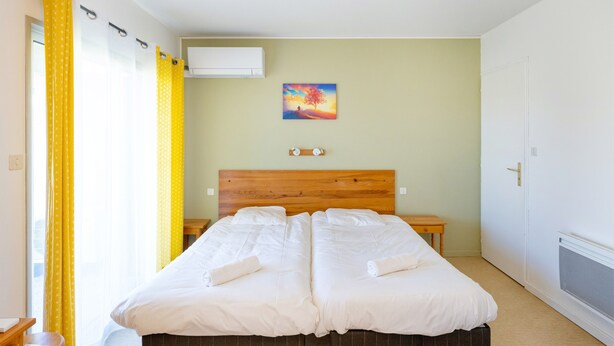 A double bed with white linens, a wooden headboard, a small bedside table, a wall-mounted air conditioner, and a painting of a tree. - Cosy Studio with Balcony, Air Conditioning & Pool Table – Cazaubon (Cazaubon)