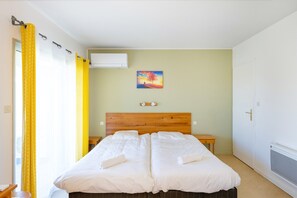 Un lit double avec du linge de lit blanc, une tête de lit en bois, une petite table de chevet, un climatiseur fixé au mur et un tableau représentant un arbre.