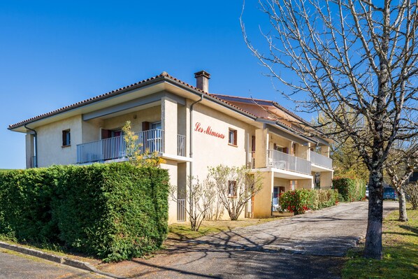 Un bâtiment à deux étages doté de balcons, une enseigne indiquant « La Mimosa », et un jardin bien entretenu.