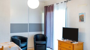 A room with a TV on a wooden dresser, a chair, a table with a patterned tablecloth, and a sliding door. - Cosy Studio with Balcony, Air Conditioning & Pool Table – Cazaubon (Cazaubon)