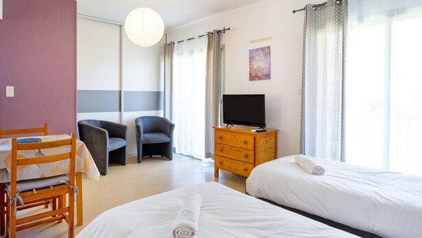 A hotel room with two beds, a desk with a chair, a TV on a wooden dresser, and a large window with curtains. - Cosy Studio with Balcony, Air Conditioning & Pool Table – Cazaubon (Cazaubon)