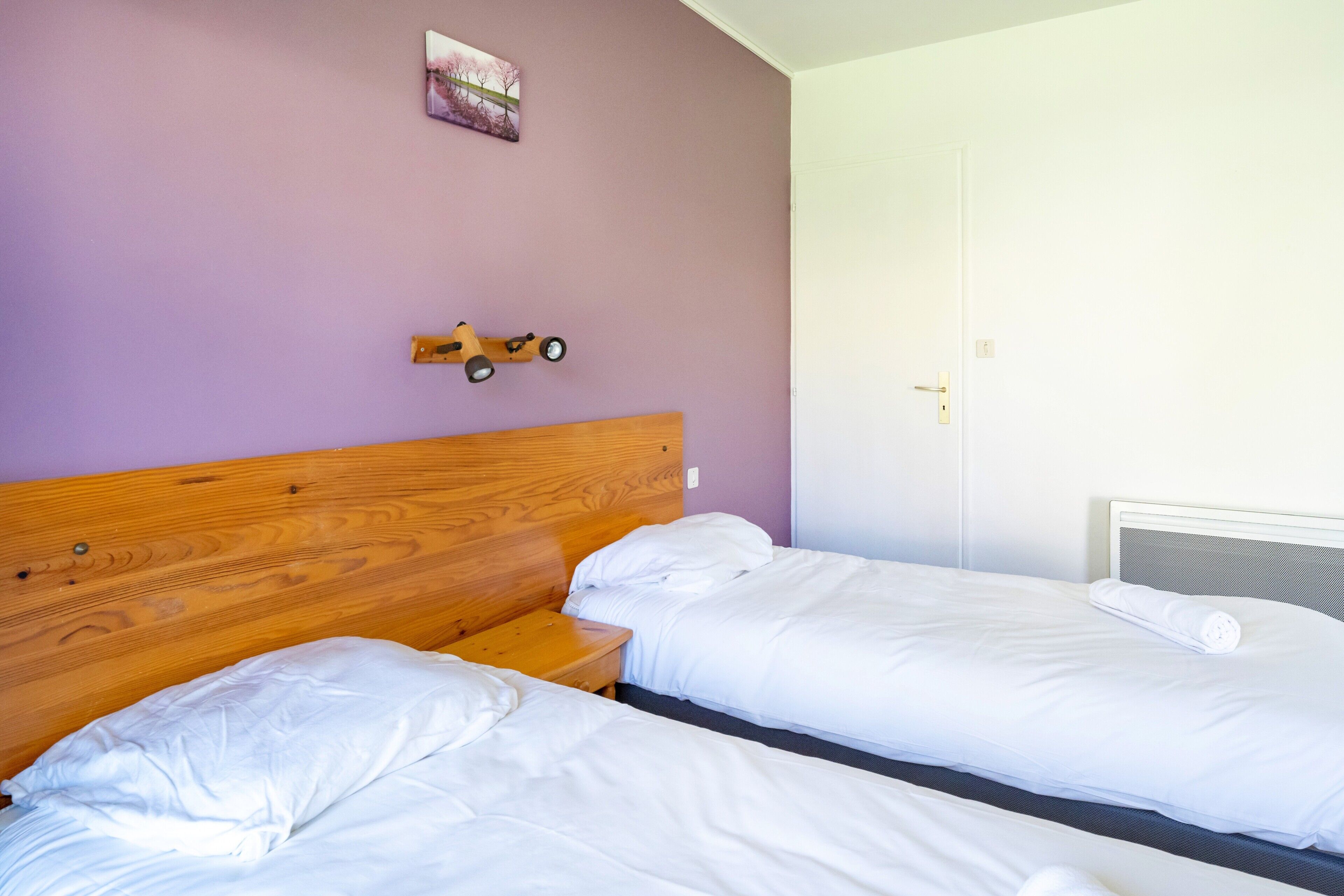 Quarto com duas camas, cabeceira de madeira, uma mesinha pequena, um brinquedo fixado na parede e uma porta.