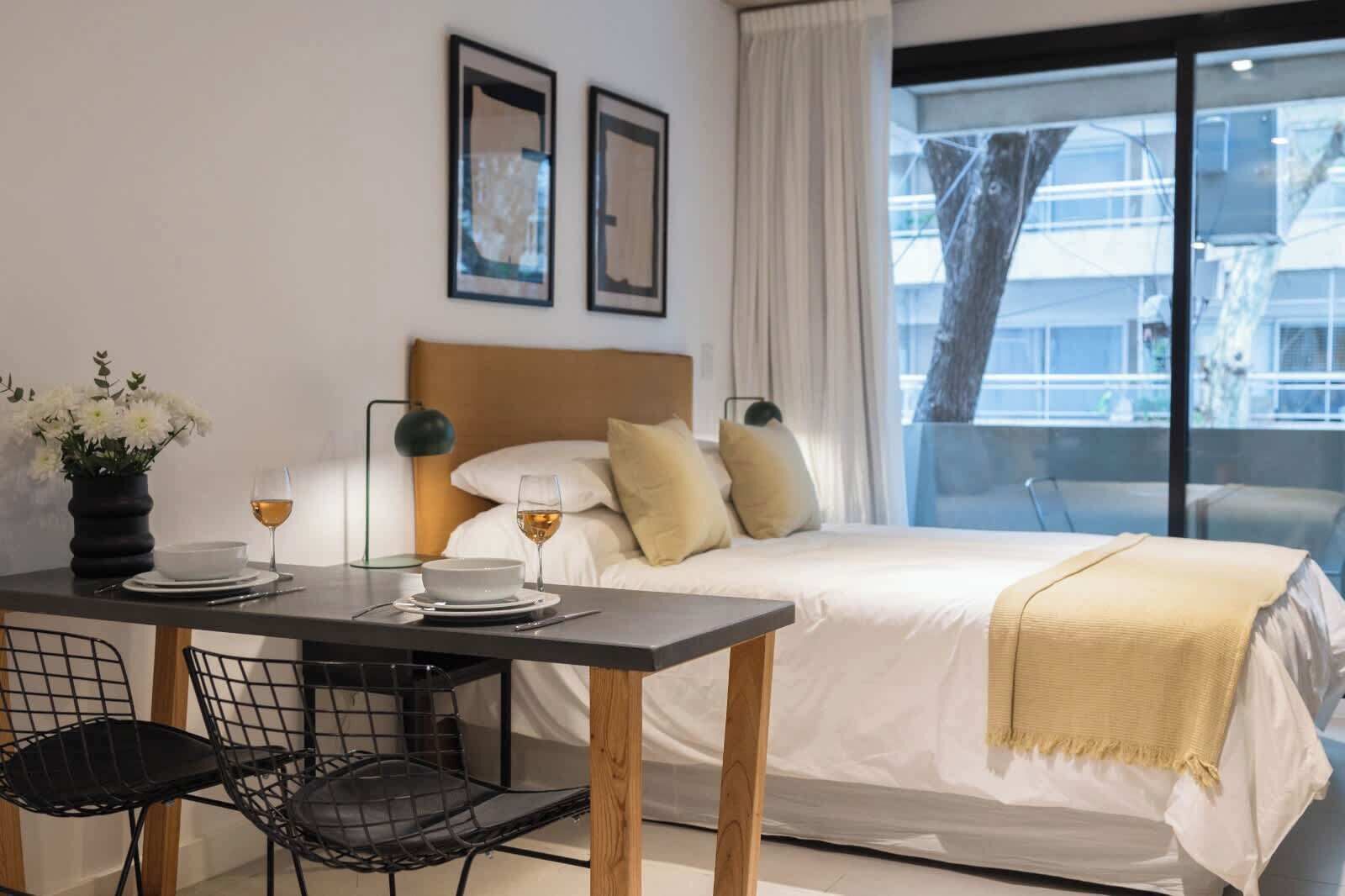 Ein Hotelzimmer mit einem Bett, einem Esstisch mit Stühlen, einer Vase mit Blumen und zwei gerahmten Bildern an der Wand.