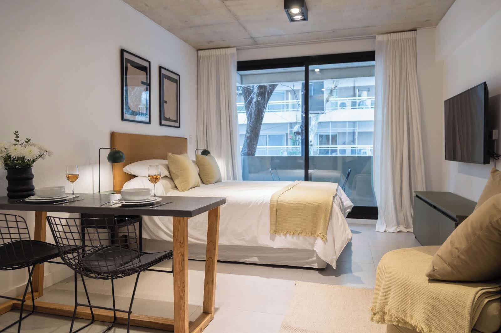 Ein Hotelzimmer mit einem Bett, einem Esstisch mit Stühlen, einem Fernseher und einem Fenster mit Vorhängen.
