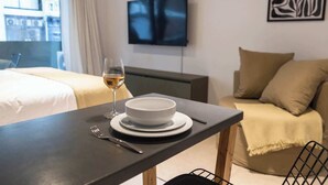 Habitación de hotel con una mesa de comedor dispuesta con un bol, plato, tenedor y una copa de vino. Un televisor está montado en la pared, y un sofá se ve al fondo.