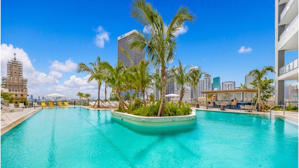 A swimming pool with palm trees, a city skyline, and a modern building.