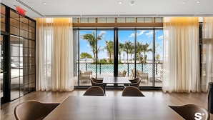A modern dining area with a large table and chairs, large windows with curtains, and a view of palm trees and a pool.