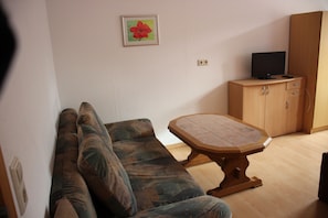 Ein Wohnzimmer mit einer Couch, einem Holztisch, einem Fernseher auf einer Kommode und einem gerahmten Bild an der Wand.