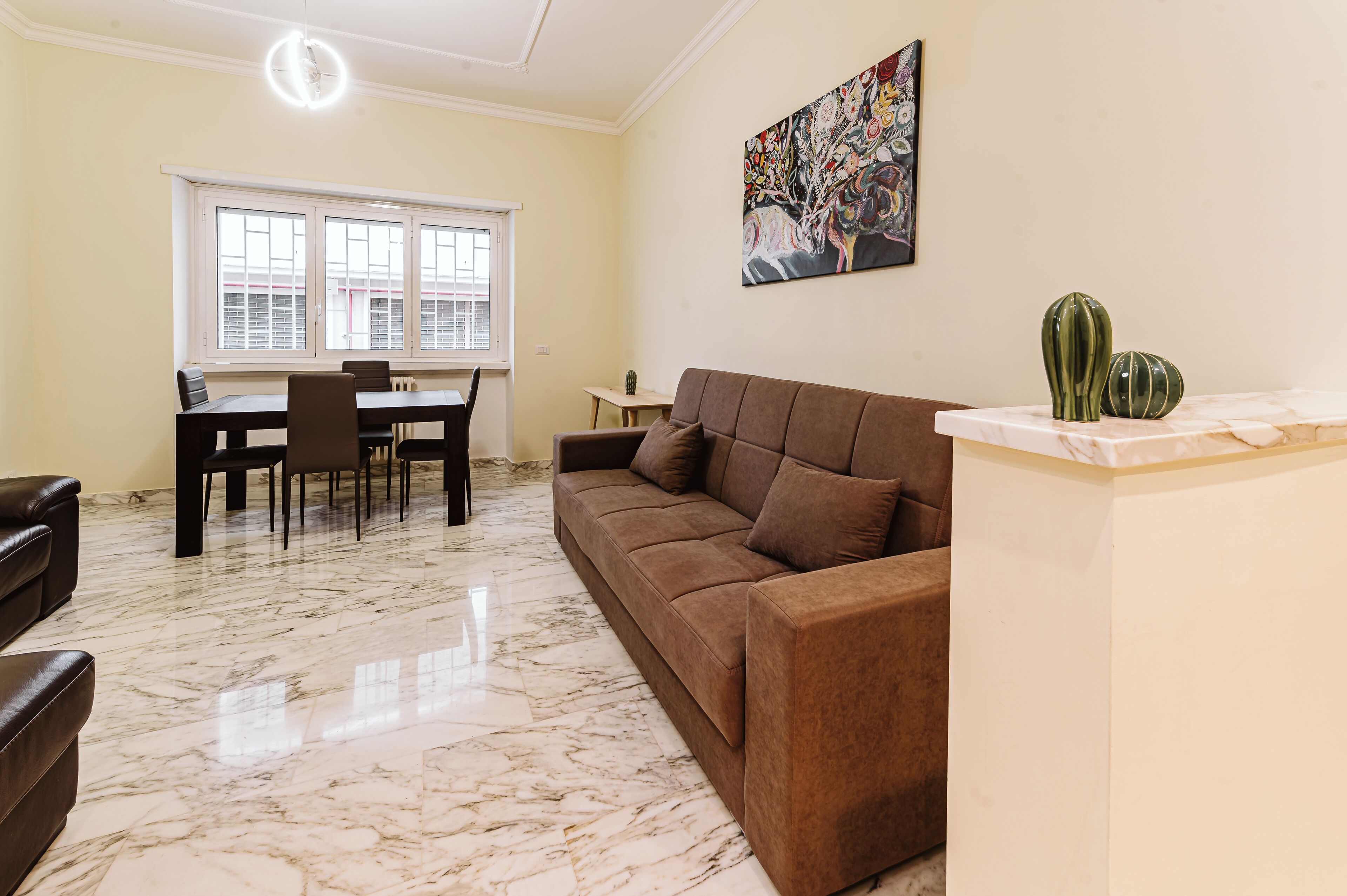 Un salon avec un canapé marron, une table à manger avec des chaises, un sol en marbre et une fenêtre avec des stores.