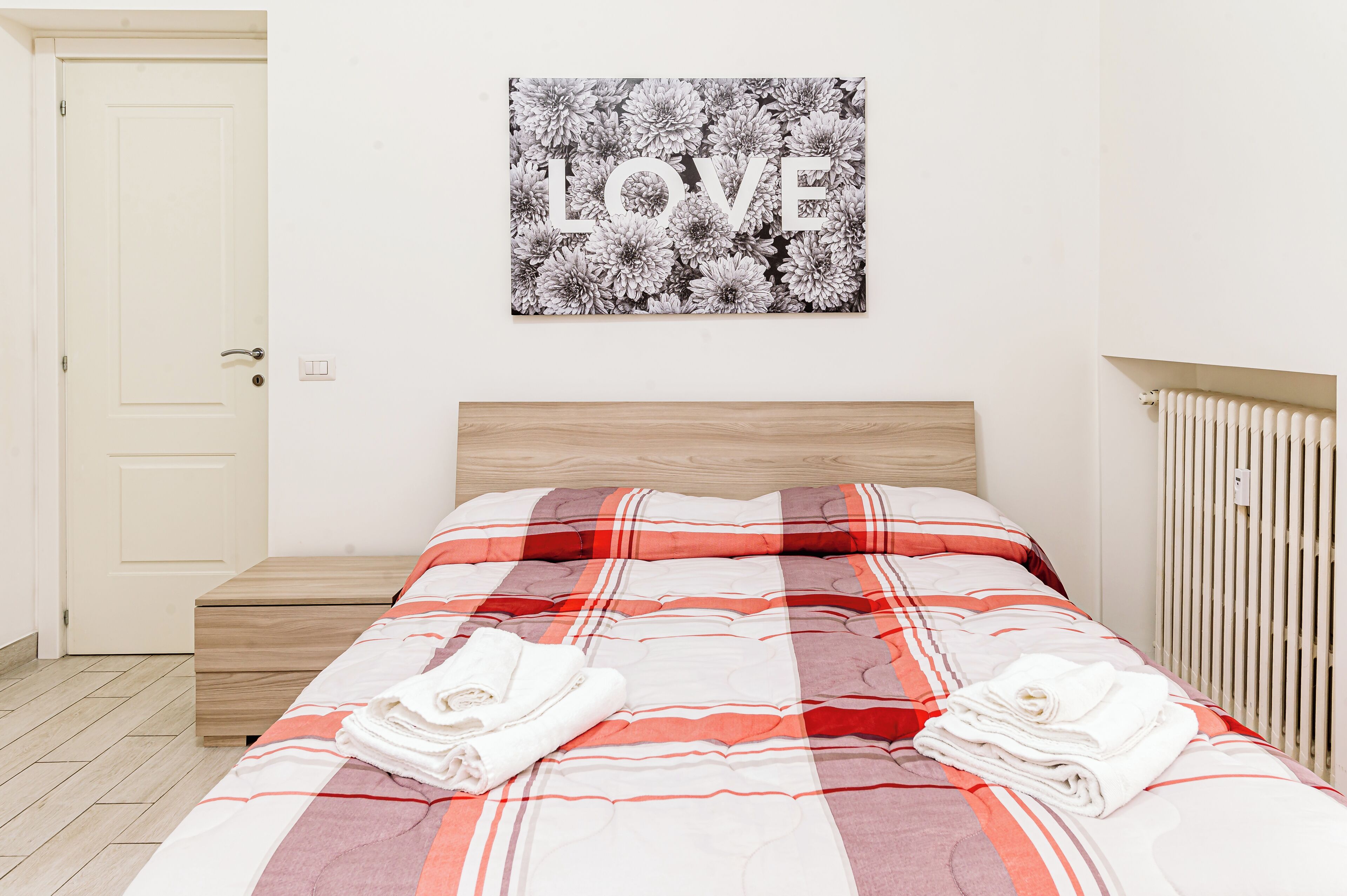 A neatly made bed with a red and white striped blanket, a wooden headboard, and a wall-mounted picture of flowers with the word "LOVE".