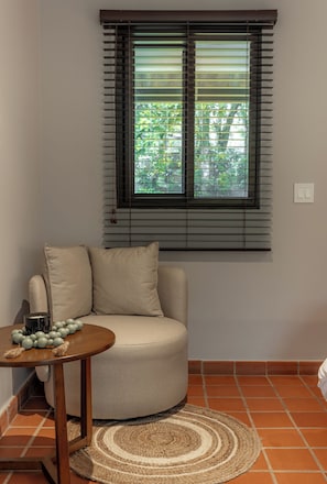 A corner room with a beige sofa, a small round table with decorative items, a window with blinds, and a tiled floor. - Hotel Alto Monte (Chiguirí Arriba)