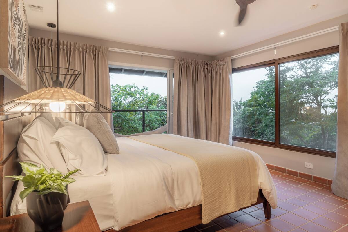 Une chambre à coucher avec un lit, des rideaux, une plante en pot et une vue sur les arbres, visible à travers de grandes fenêtres.