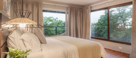 Ein Schlafzimmer mit einem Bett, Vorhängen, einer Topfpflanze und Blick auf Bäume durch große Fenster.