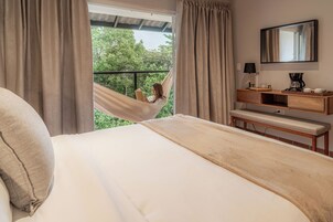 A bedroom with a bed, a hammock, a bedside table with a lamp, and a view of greenery outside.