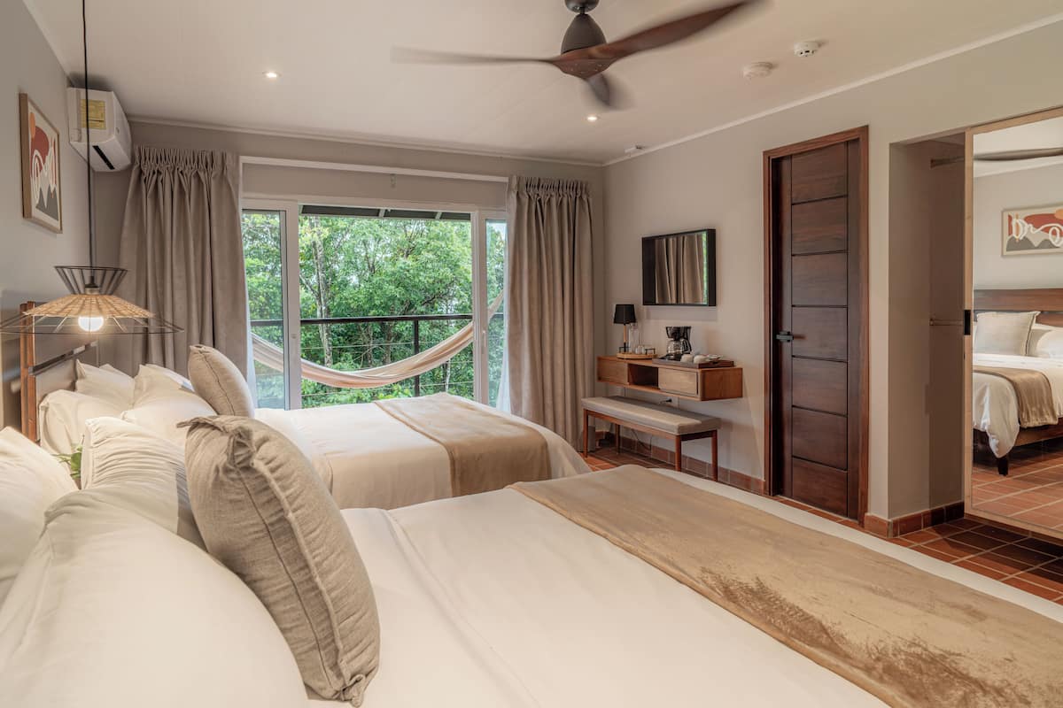 Une chambre avec deux lits, un hamac, un ventilateur de plafond, une fenêtre avec des rideaux et une porte donnant sur une autre pièce.