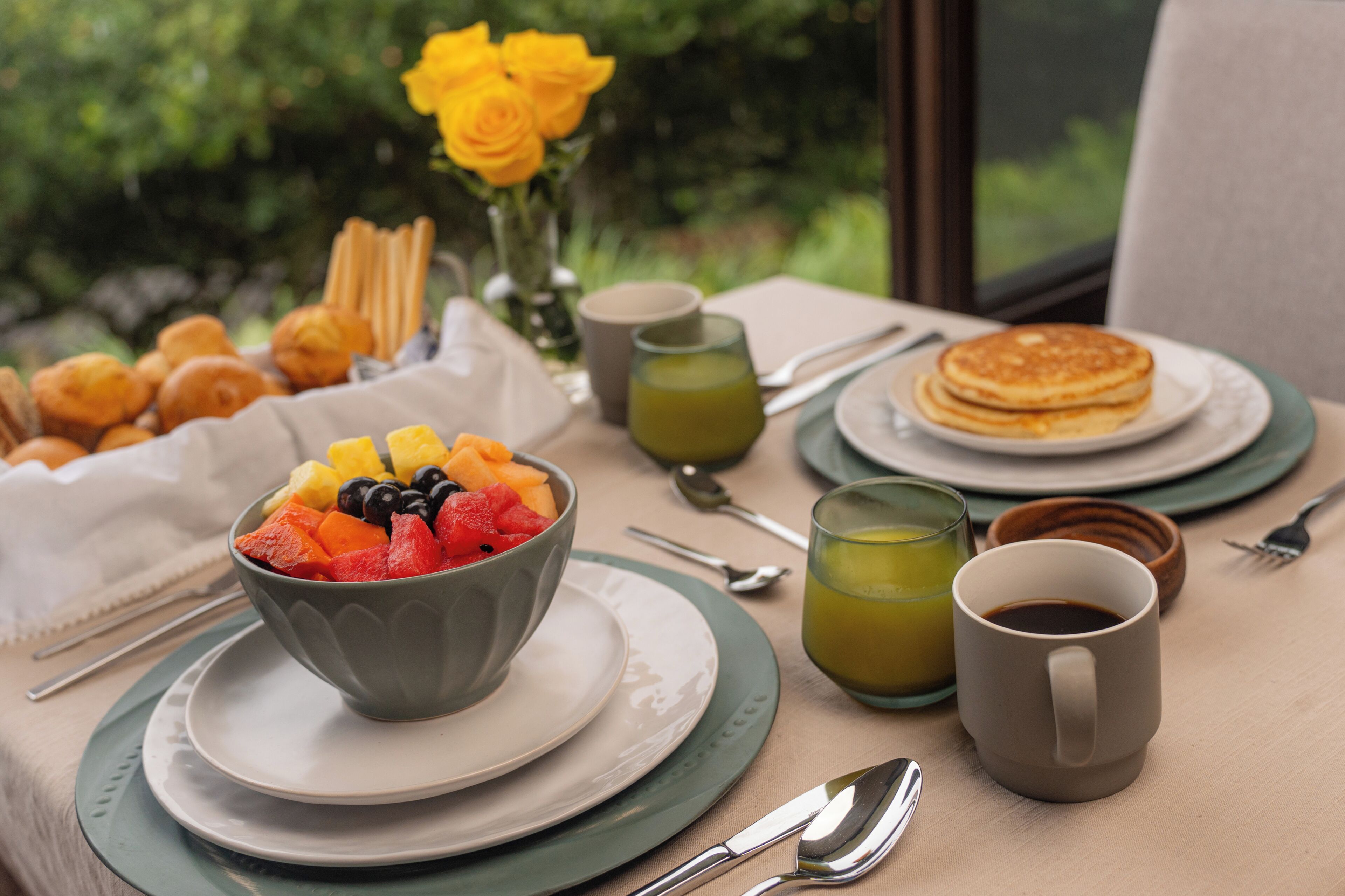 Un desayuno con un tazón de fruta, panqueques y café sobre una mesa.