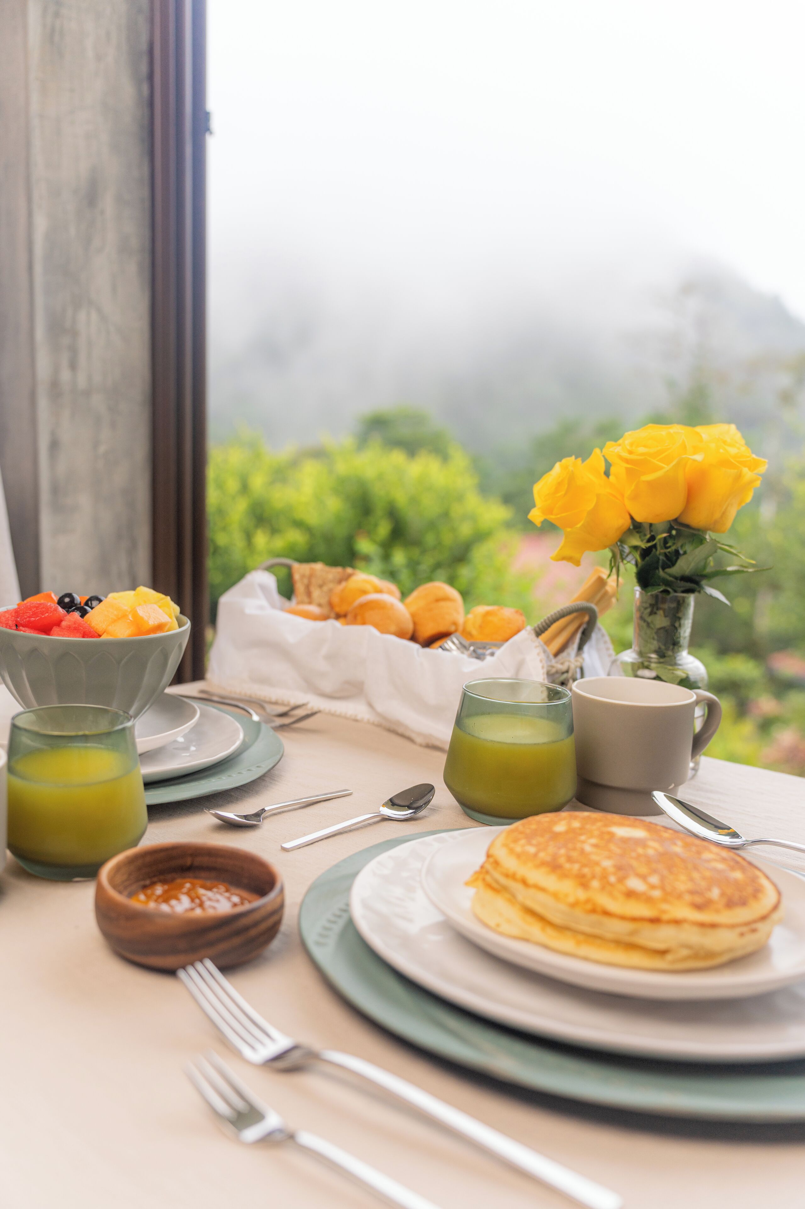 Una mesa de desayuno con panqueques, fruta y un ramo de rosas amarillas.