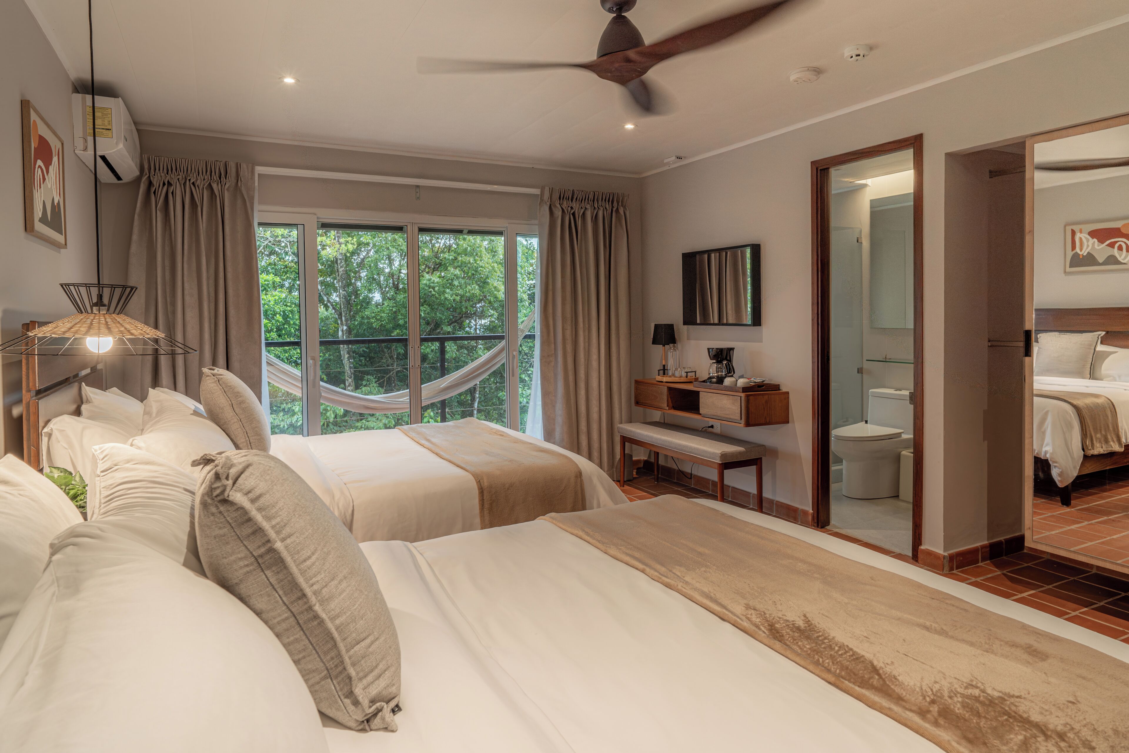 A bedroom with two beds, a ceiling fan, a nightstand, a desk, a chair, a window with curtains, and a view of greenery outside.