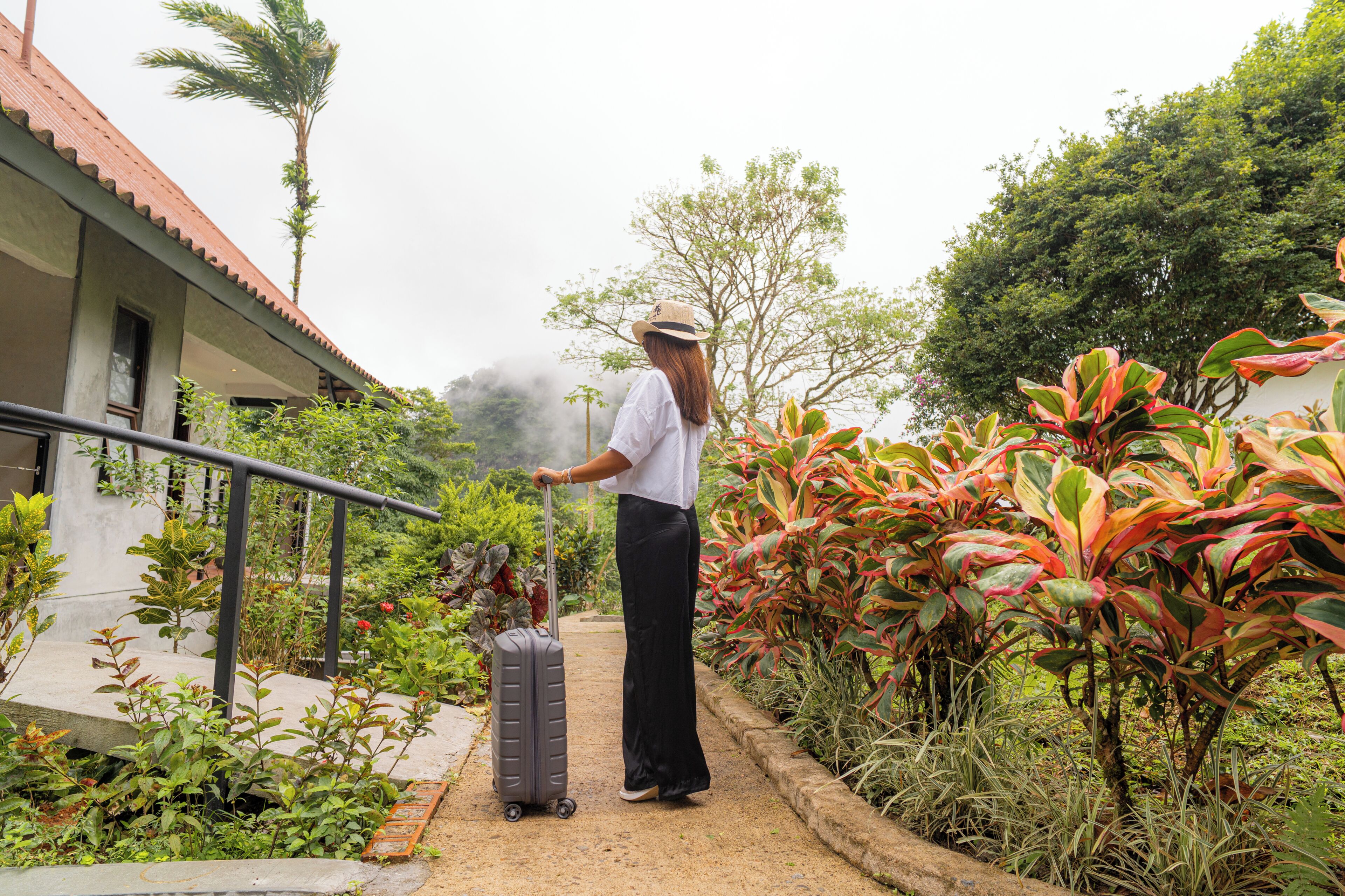 A person with a suitcase on a pathway surrounded by lush greenery and a building with a red roof.