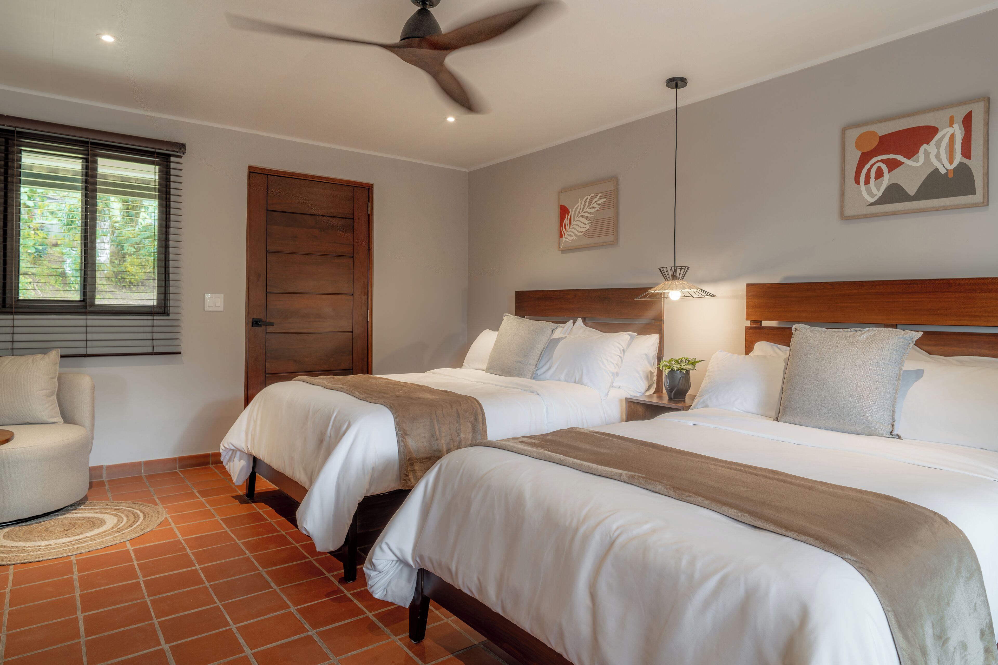A bedroom with two beds, a ceiling fan, a window with blinds, and a painting on the wall.