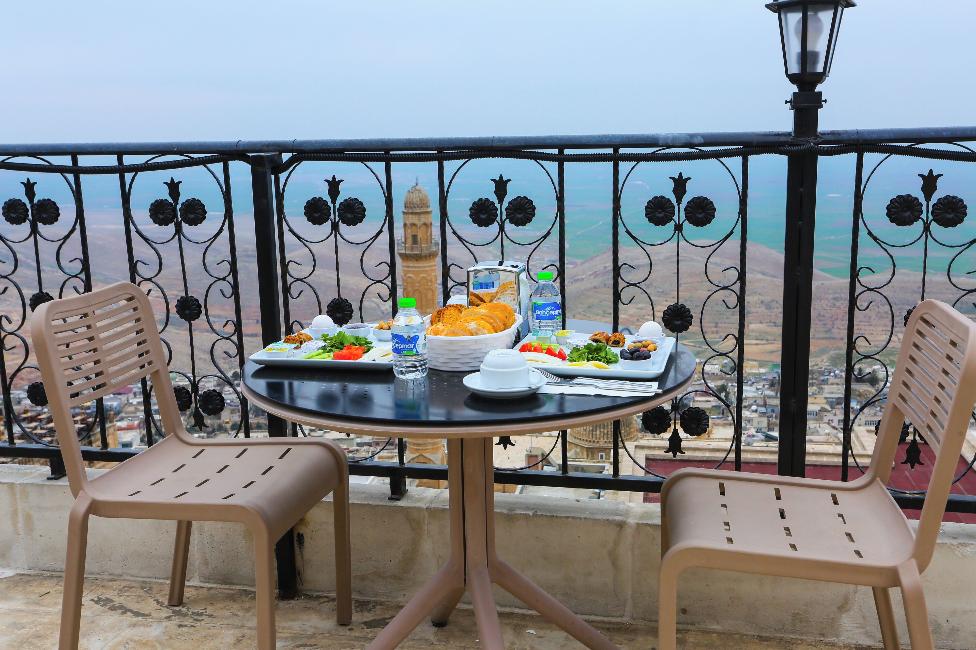 A balcony with a table set for a meal, two chairs, a lamp, and a view of a distant building.