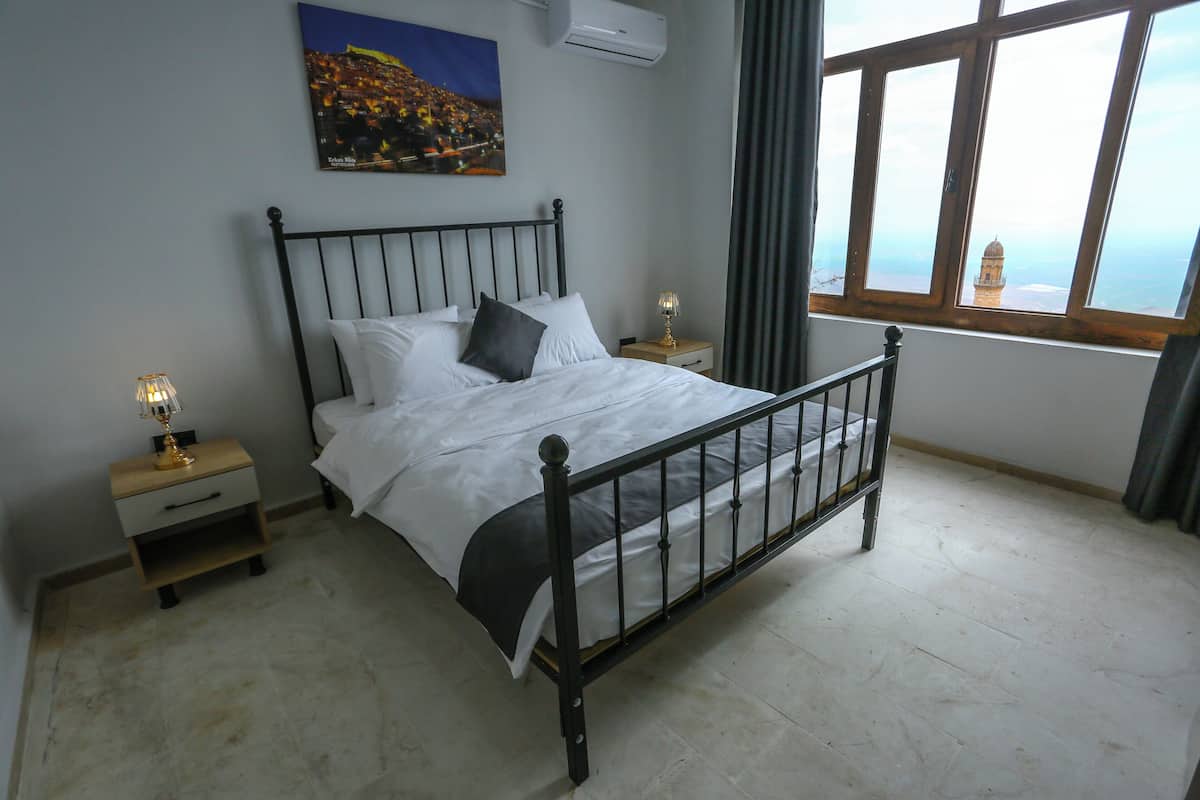 Un dormitorio con cama, mesitas de noche, lámpara, un cuadro y ventana con vistas.