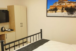 Una cama con cabecera negra, un armario de madera, un televisor de pantalla plana y un cuadro enmarcado de un edificio en la pared.