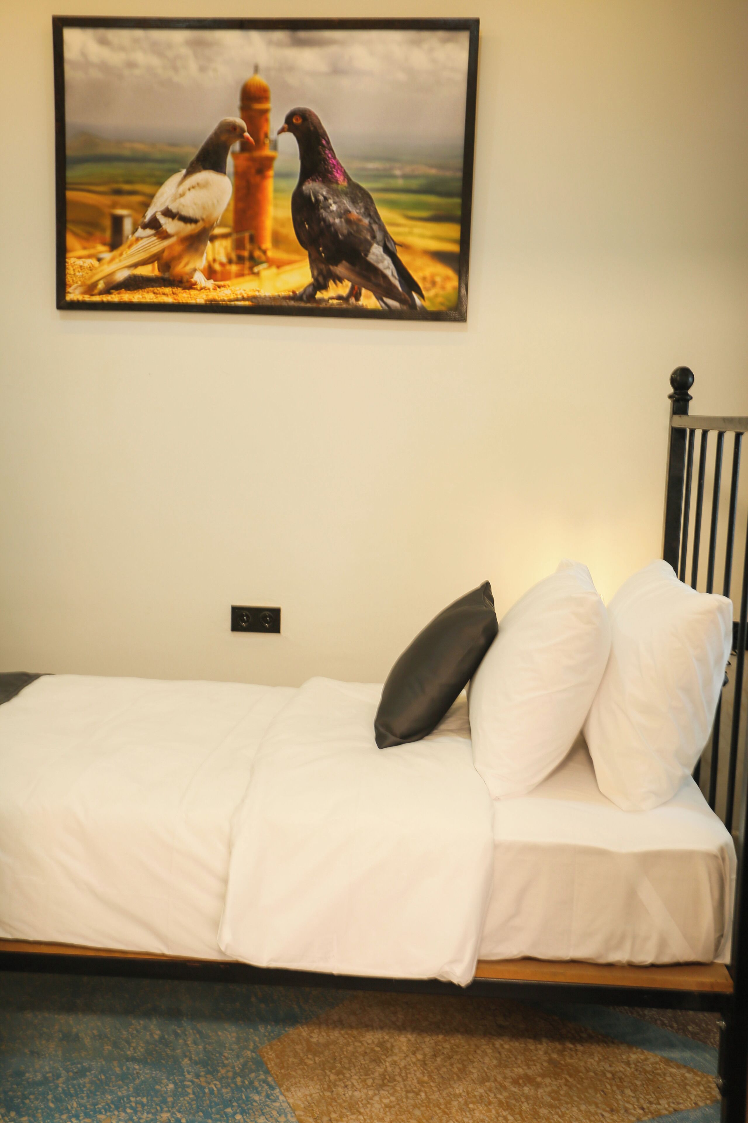 Ein Bett mit weißer Bettwäsche und einem schwarzen Kopfkissen, ein Gemälde von Tauben an der Wand und eine Steckdose.