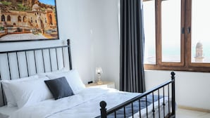 Een bed met witte lakens, een nachtkastje met een lamp, een raam met uitzicht op een gebouw, en een schilderij aan de muur.