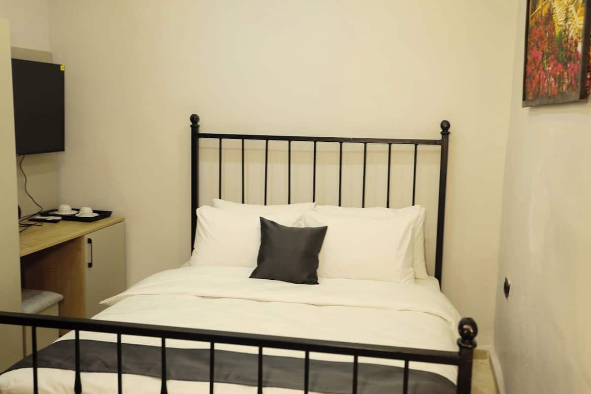 Una cama individual con estructura metálica negra, ropa de cama blanca y almohada negra. Hay un escritorio con una silla y un televisor de pantalla plana montado en la pared.