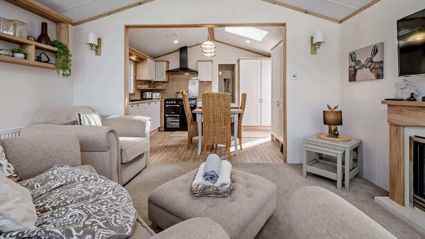 Un espacio de sala acogedor con sofá, reposapiés y una mesita con lámpara. Se observa una cocina al fondo con una mesa y sillas de comedor.