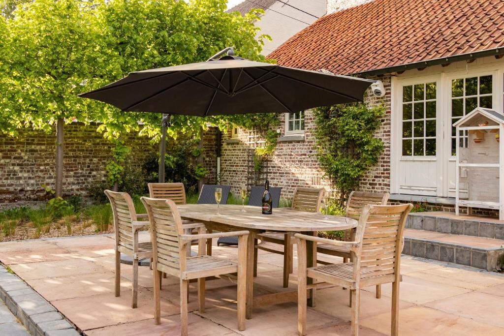Een terras met een ronde houten tafel en stoelen, een parasol en een bakstenen muur.