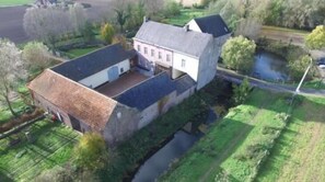 Une maison à deux étages avec une grande véranda à l’avant, un petit garage attenant et une rivière en arrière-plan.