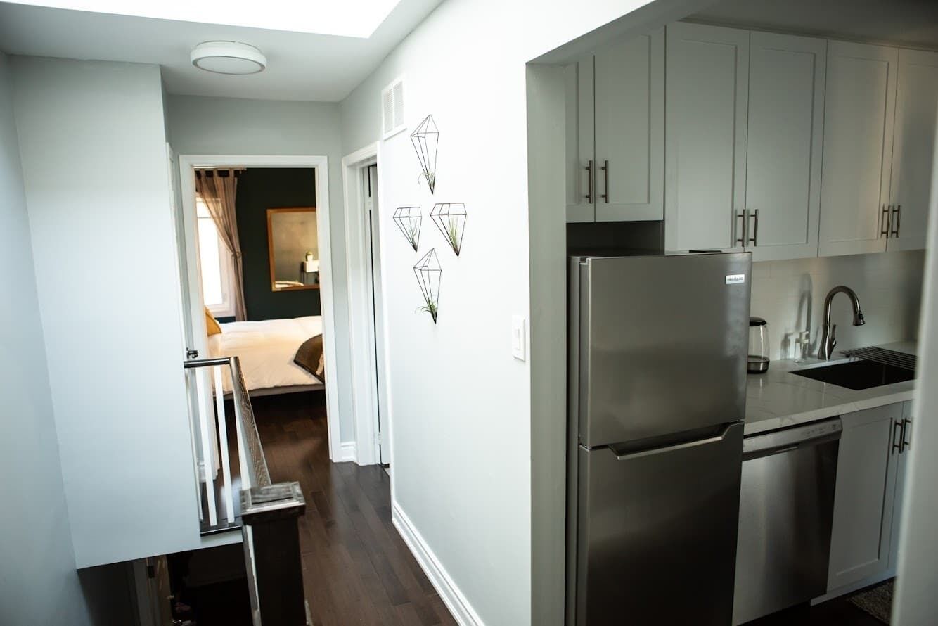 A modern kitchen with stainless steel appliances, white cabinetry, and a small dining area with a bed visible through an open door.