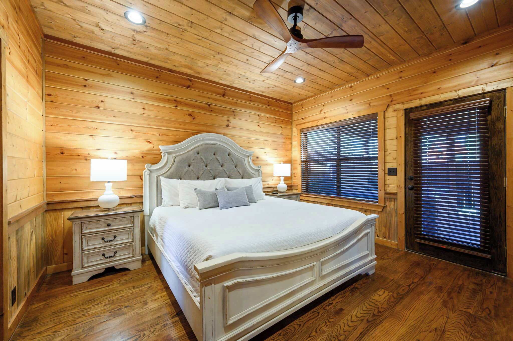 Ein Schlafzimmer mit Holzwandverkleidung, einem großen Bett, zwei Nachttischen mit Lampen, einem Deckenventilator und einem Fenster mit Jalousien.