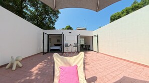 Ein Zimmer mit einem Bett, einem kleinen Tisch und einem Stuhl. Ein großes Fenster und eine Tür führen nach draußen.