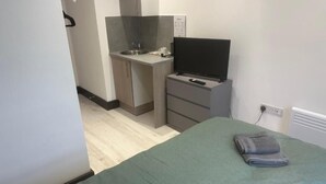 Un dormitorio con cama, televisor en un mueble, pequeña cocina americana con lavabo y microondas, y un enchufe en la pared.