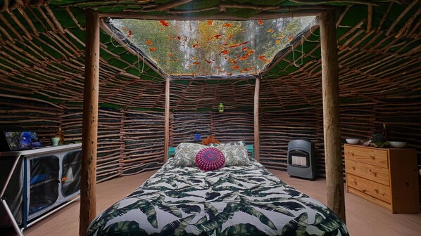 A room with a bed, a small table, a speaker, and a wall with a naturalistic design.