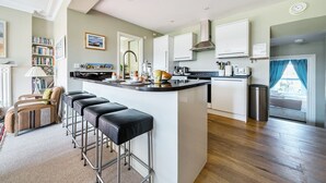 A modern kitchen with a central island, bar stools, and a dining area with a sofa and bookshelf. - 3 Monteagle - sleeps 6 guests  in 3 bedrooms (Wells-next-the-Sea)