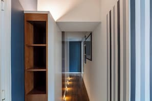 Ein schmaler Korridor mit einem Holzregal links, einer blauen Wand rechts und einer Reihe gerahmter Bilder an der Wand.