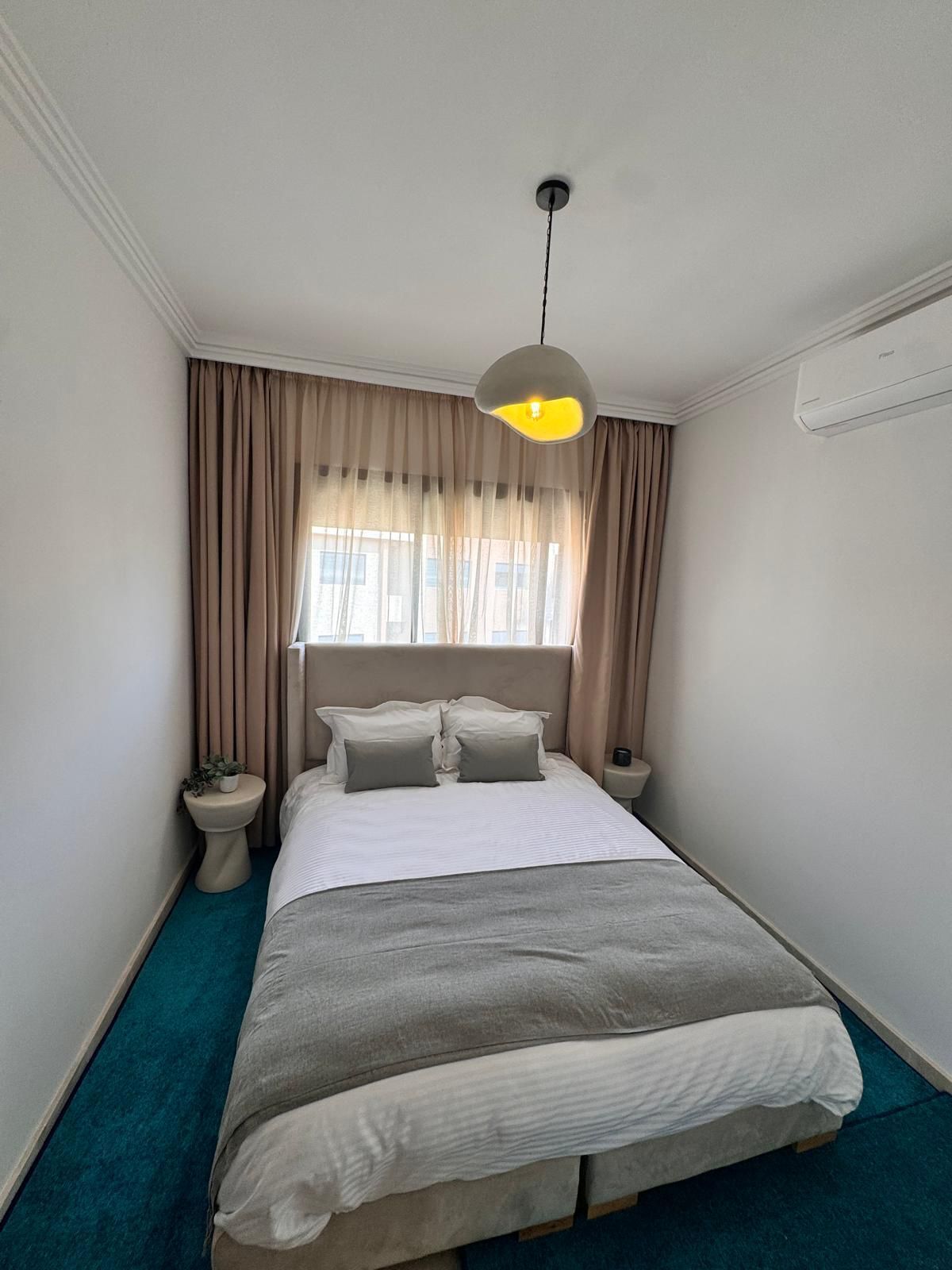 Une chambre à coucher comprenant un lit, une table de chevet, une commode, un luminaire suspendu et une fenêtre avec des rideaux.