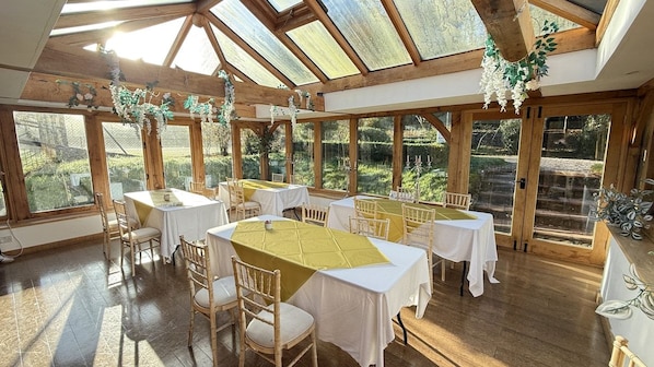 A spacious room with multiple tables and chairs, large windows, and a wooden ceiling structure.