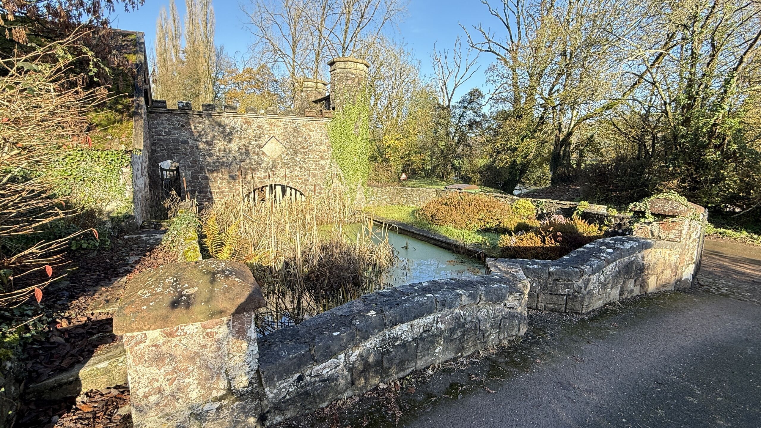 Een stenen muur met een poort, een kleine vijver en omliggende beplanting.